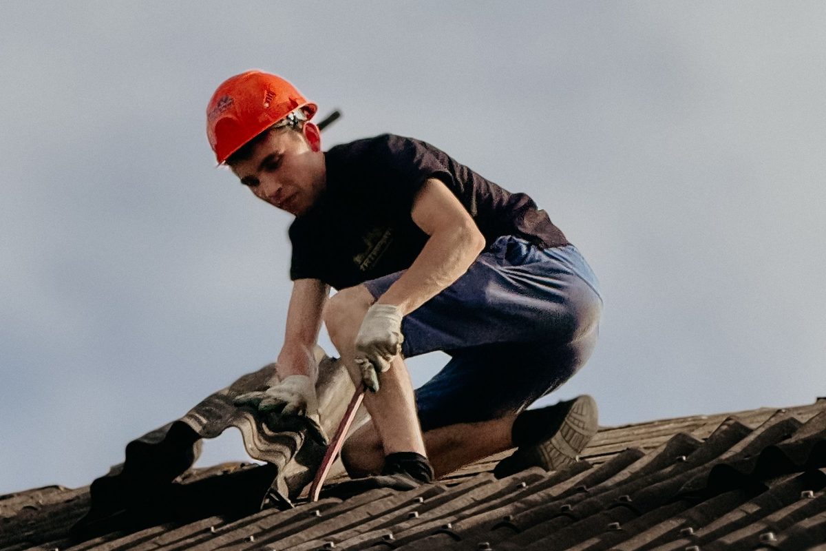 A roofer in an orange hard hat kneeling on a corrugated roof, lifting a roofing sheet with a pry bar to inspect underneath.